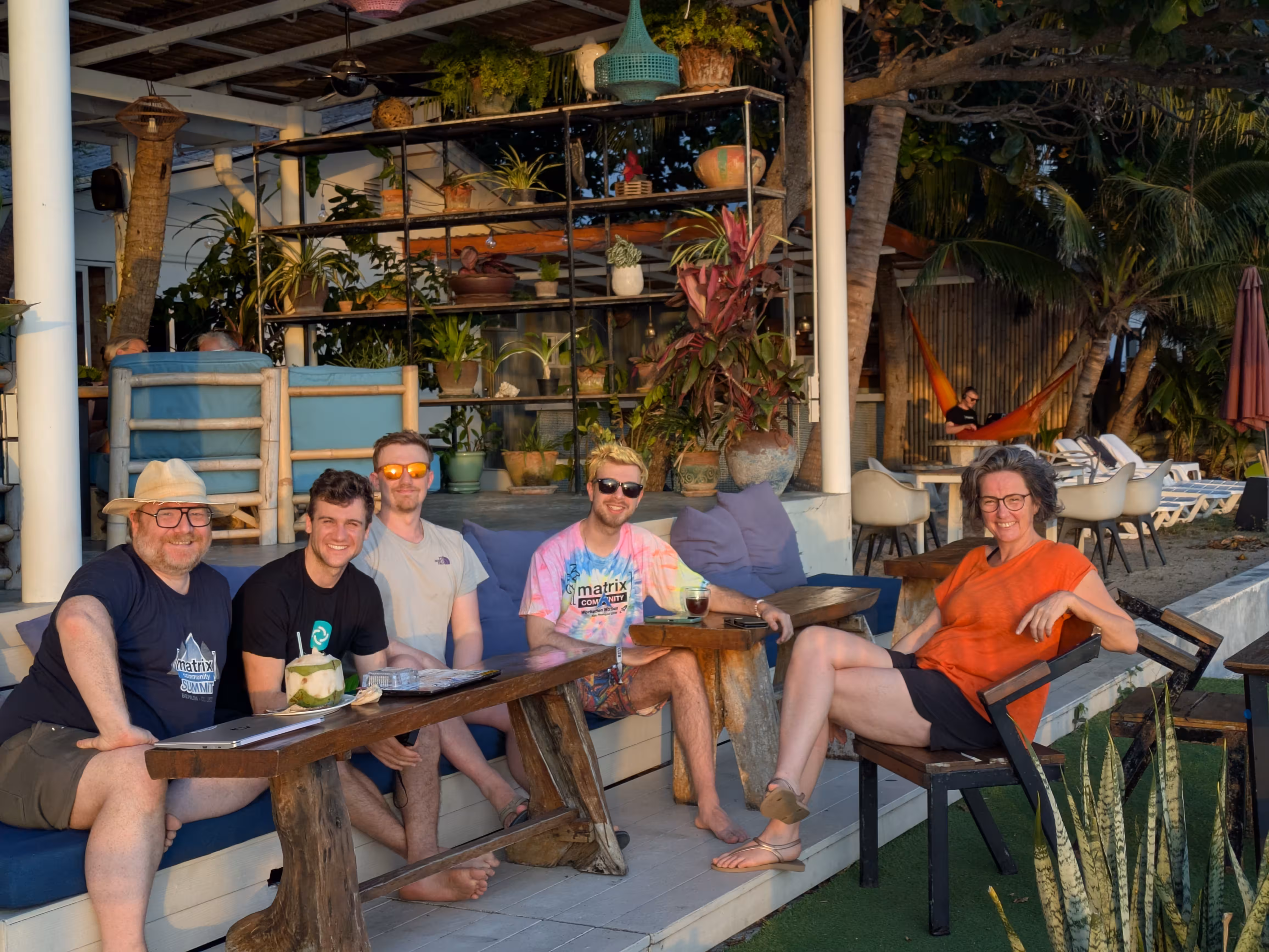A group of people in summer outfits lounging on a sofa, enjoying the sunset. One is participating in a meeting from a hammock in the background.